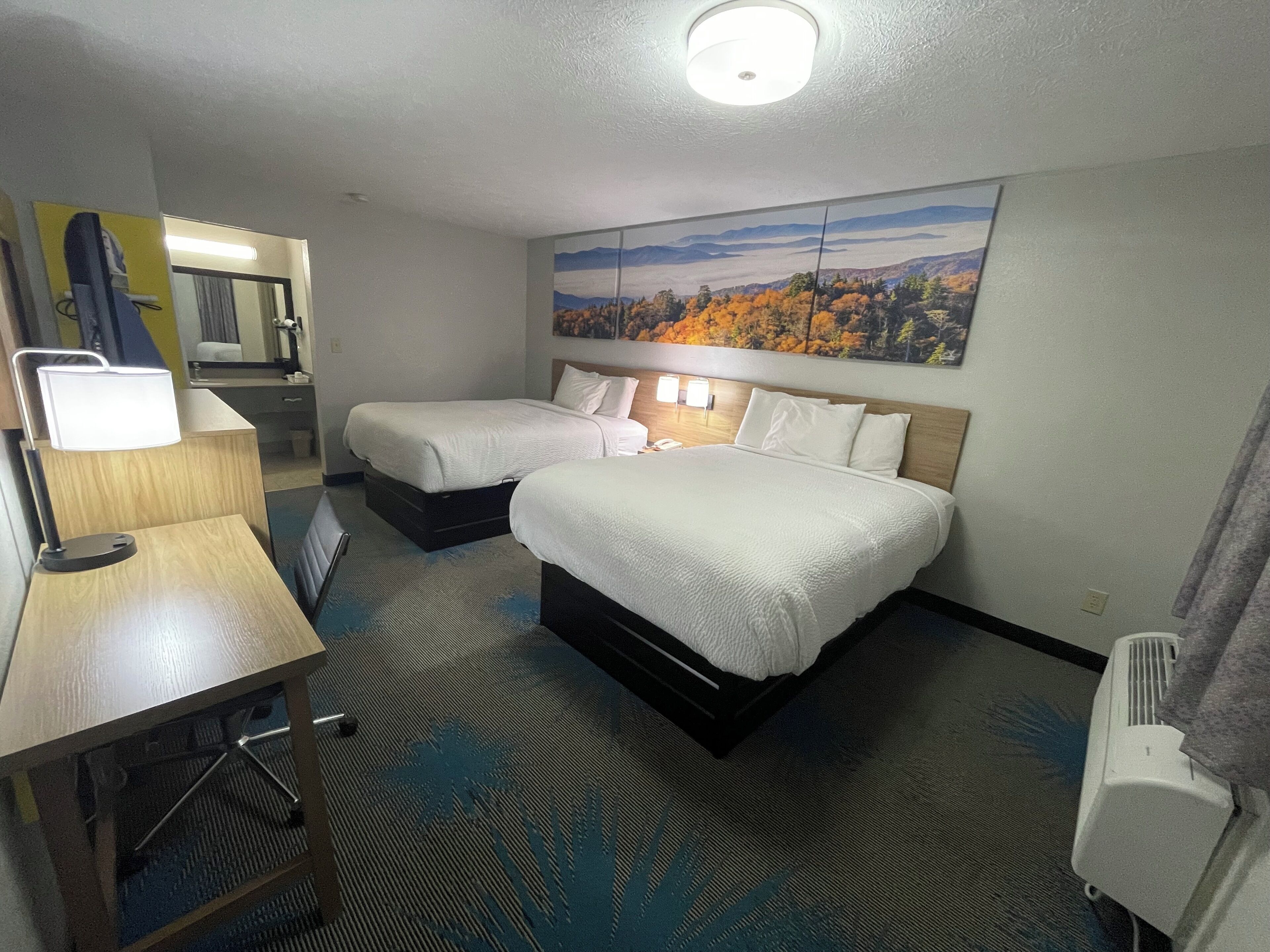 A hotel room with two beds, a desk with a lamp, a mirror, and a painting of a mountain landscape.