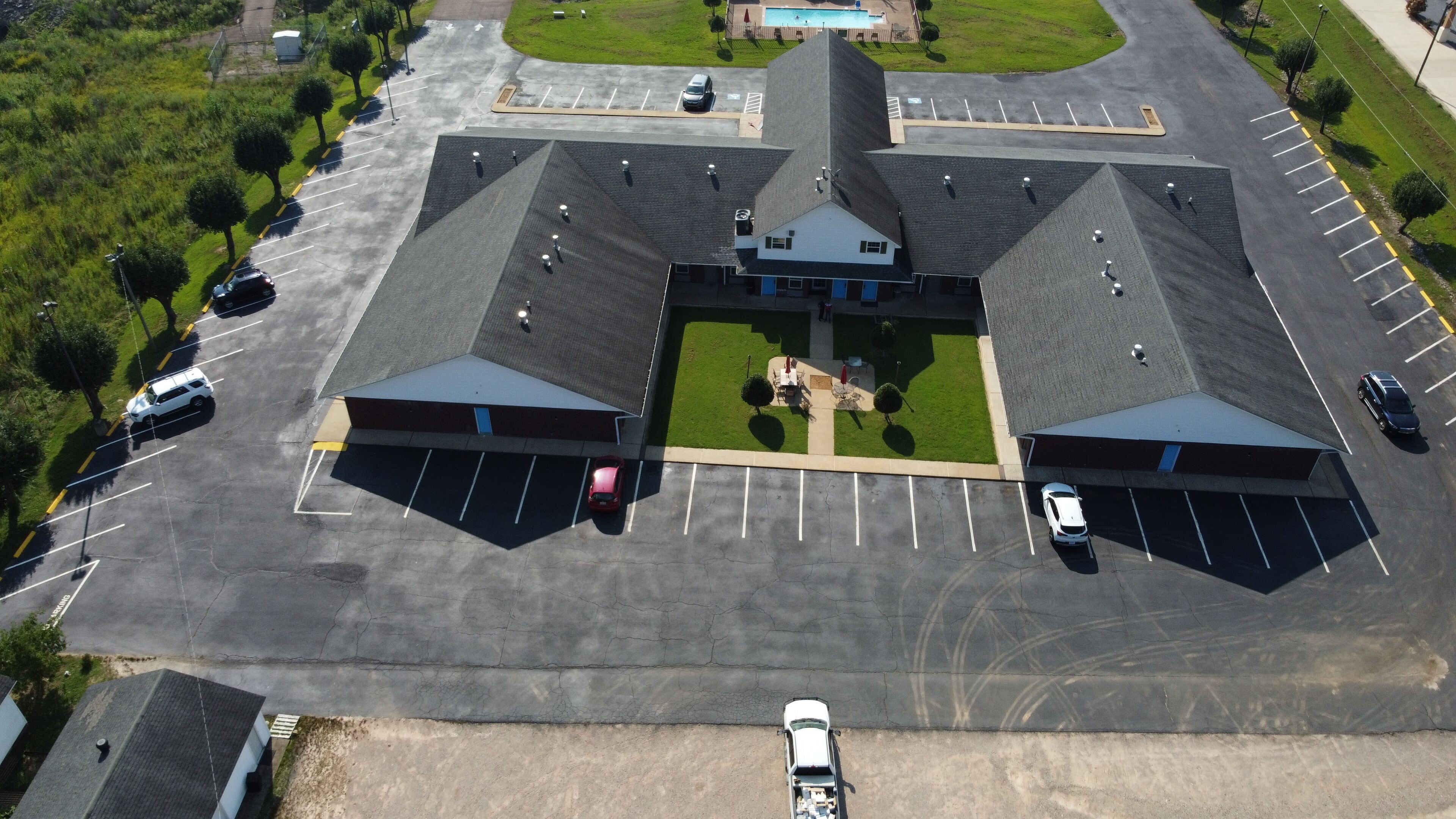 Vista aérea de un estacionamiento con varias filas de espacios, un área central ajardinada y un edificio con una estructura de techo distintiva.
