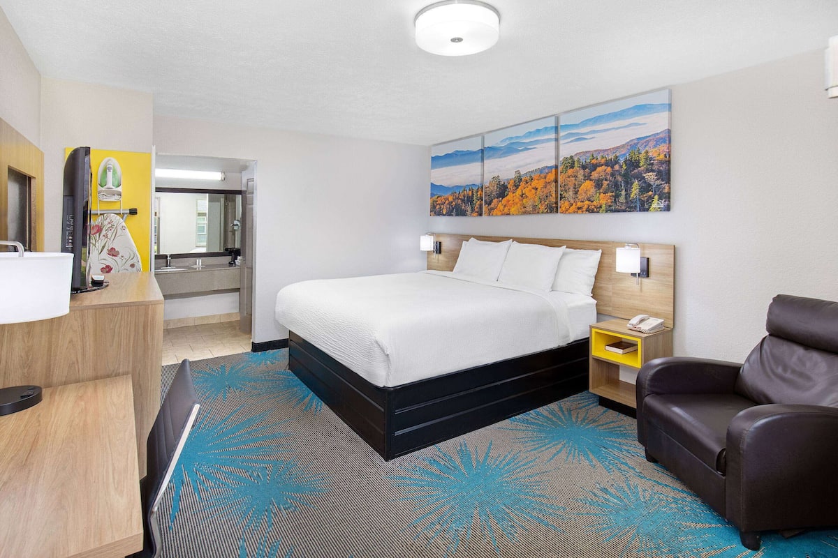 A hotel room with a bed, a desk, a chair, a nightstand, and a painting on the wall.