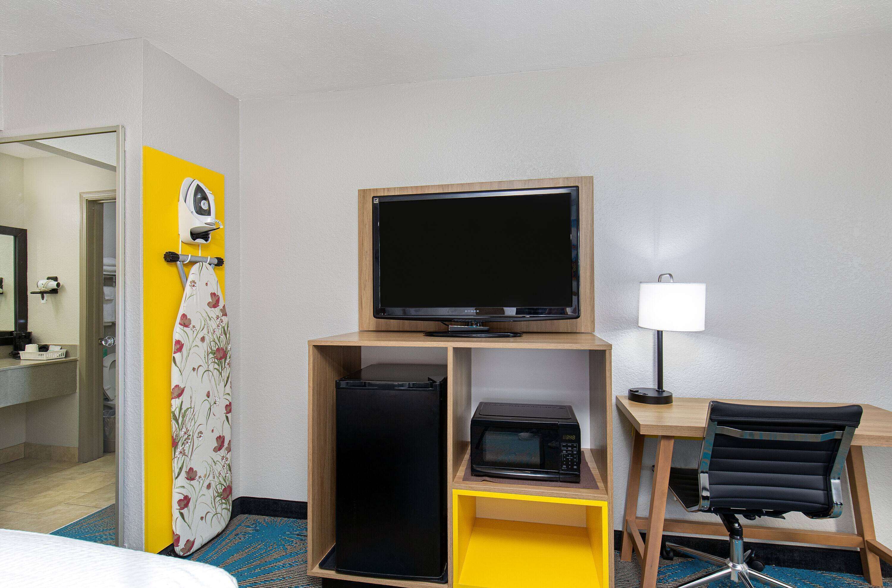 A hotel room with a bed, a desk with a computer monitor, a microwave, a refrigerator, a chair, a lamp, and a bathroom visible through an open door.