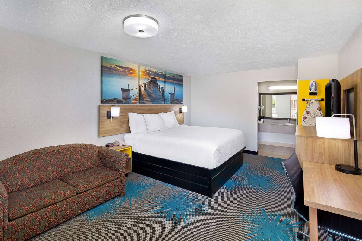 A hotel room with a bed, a sofa, a desk, and a painting of a sunset over a pier.
