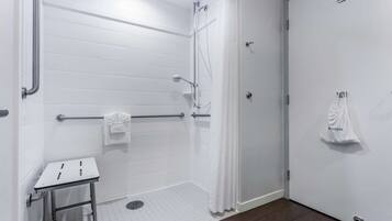 Un baño con paredes de azulejos blancos, una ducha con puerta de cristal, un asidero, una mesita pequeña y un perchero con una toalla.