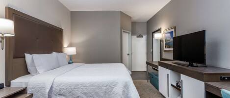 A hotel room with a bed, bedside table, lamp, TV, and a desk with a chair.
