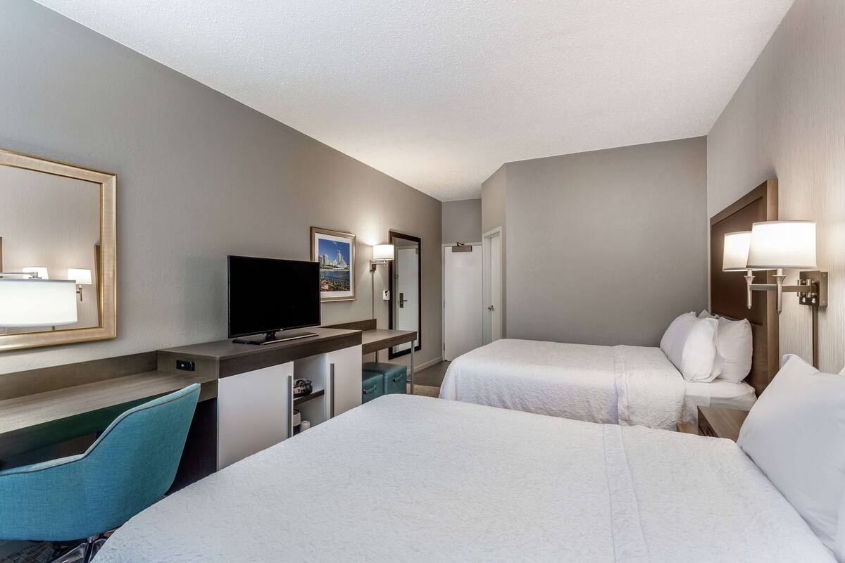 Ein Hotelzimmer mit zwei Betten, einem Schreibtisch mit Fernseher, einem Sessel und einer Schreibtischlampe.