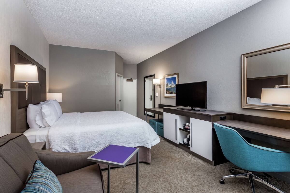 Ein Hotelzimmer mit einem Bett, einem Schreibtisch, einem Sessel, einem Fernseher und einem kleinen Tisch mit einer violetten Ordner.