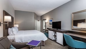 A hotel room with a bed, a desk, a chair, a TV, and a small table with a purple folder.