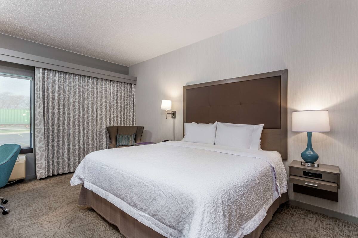 Ein Hotelzimmer mit einem großen Bett, einem Sessel, einem Schreibtisch mit Lampe und einem fenster mit Vorhängen.