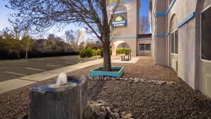 A Days Inn & Suites building with a clear sky, a parking lot, a fountain, and a tree in the foreground. - Days Inn & Suites by Wyndham Airport Albuquerque (Albuquerque)