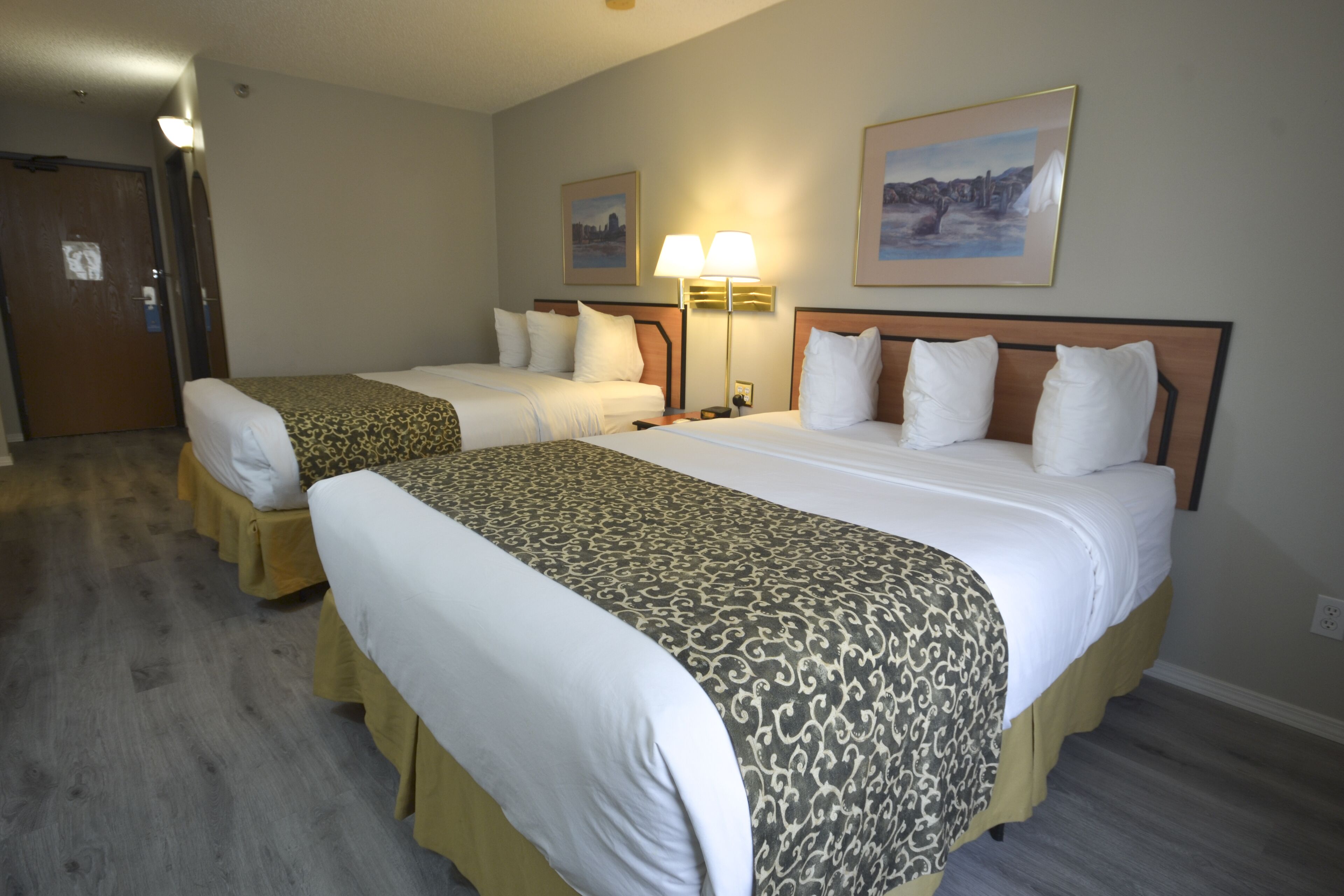 Habitación de hotel con dos camas, cabecero de madera, mesita de noche, lámpara y dos cuadros en la pared.