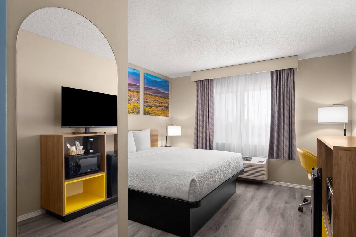 A hotel room with a bed, a TV, a desk with a lamp, a microwave, a coffee maker, and artwork on the wall.