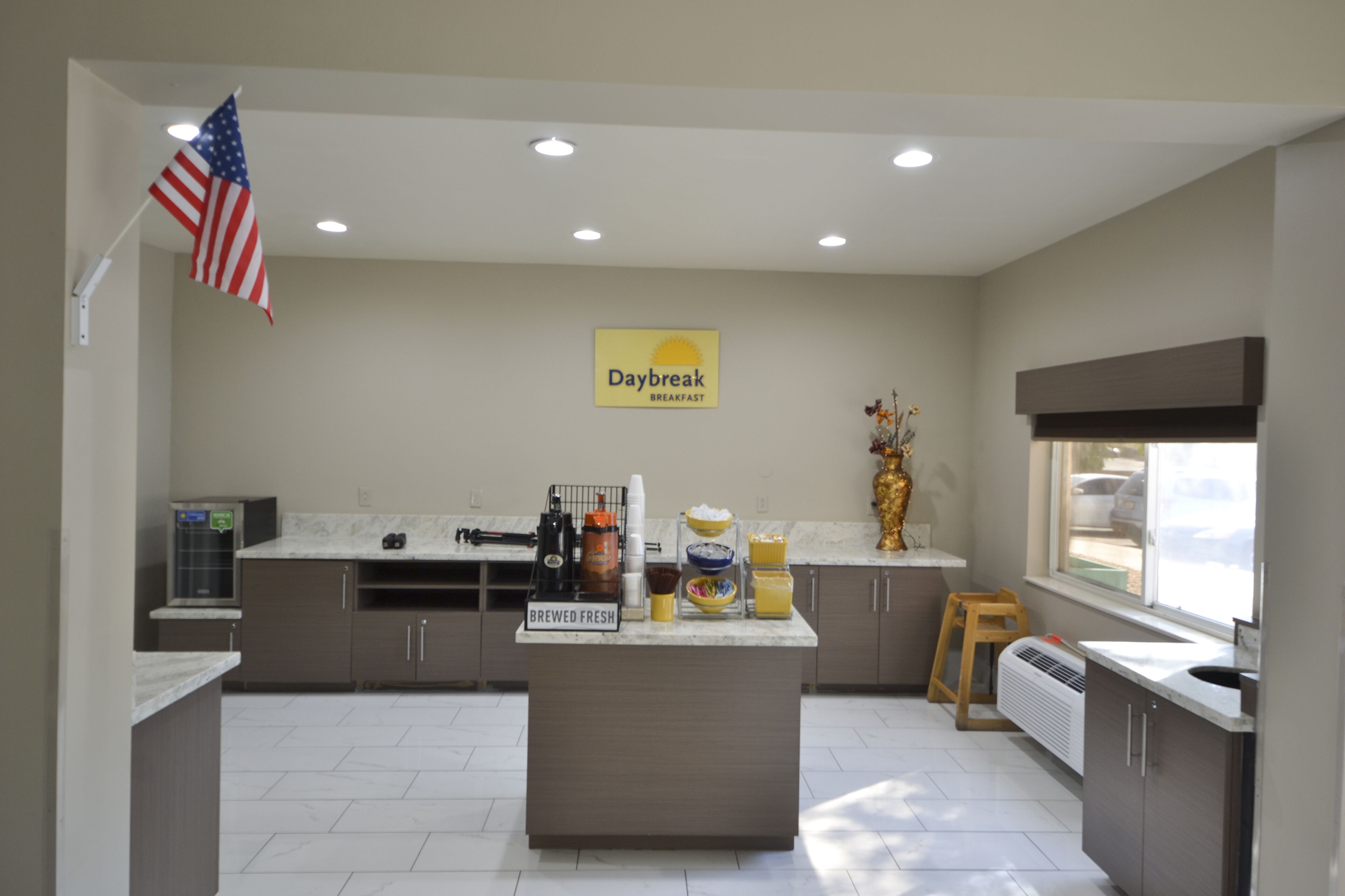 A modern kitchen with a central island labeled "BREWED FRESH," a "Daybreak BREAKFAST" sign, and an American flag.