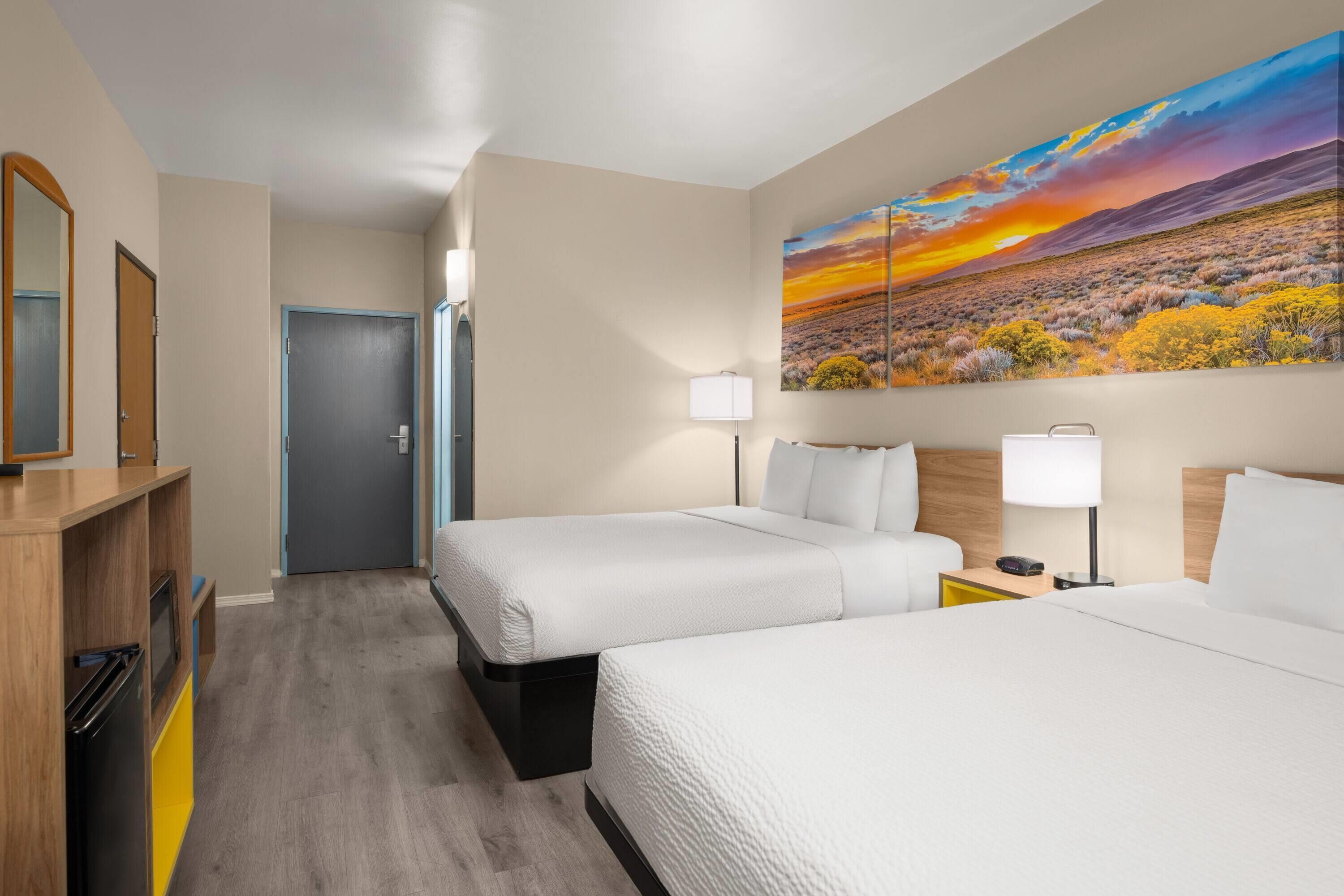 Une chambre d’hôtel avec deux lits, un tableau représentant un paysage au coucher du soleil, un bureau avec une lampe et une porte.