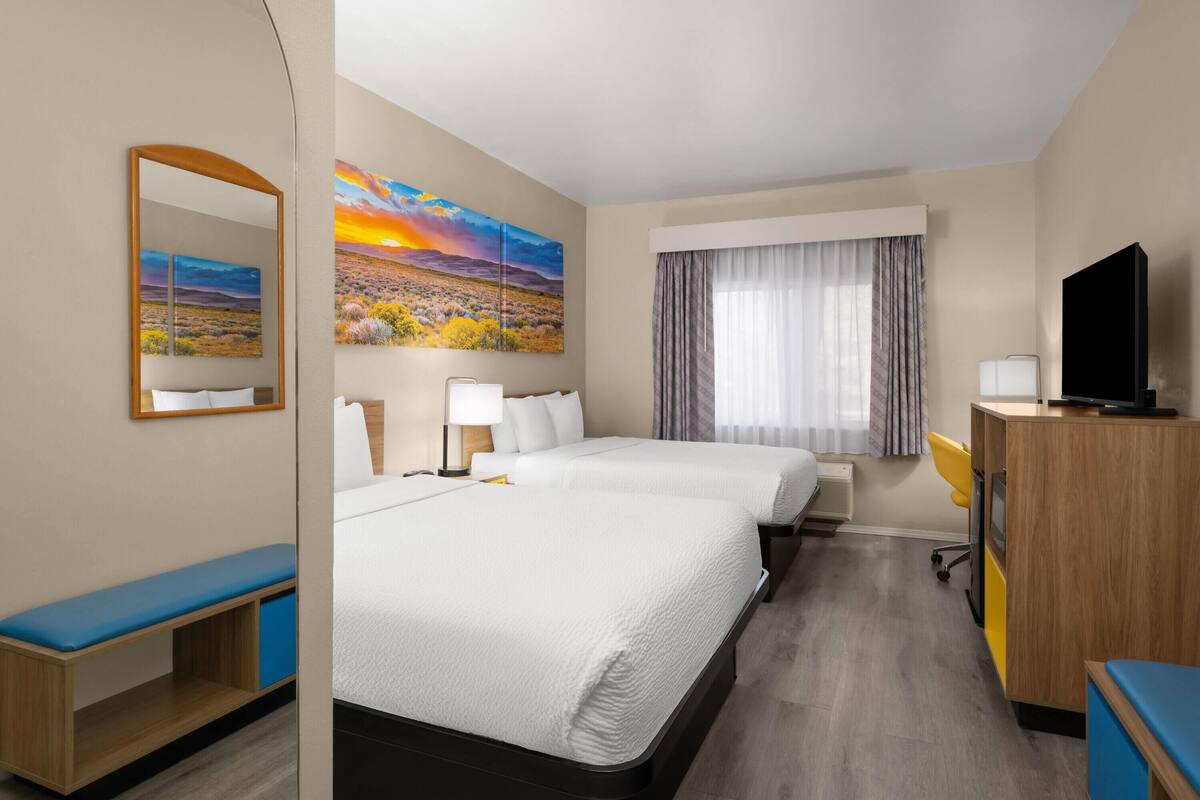 A hotel room with two beds, a desk, a TV, and a large window with curtains.