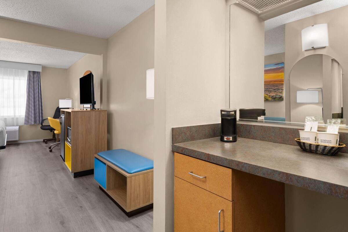 A hotel room with a kitchenette, a desk, a chair, a blue bench, a coffee machine, and a basket of amenities.