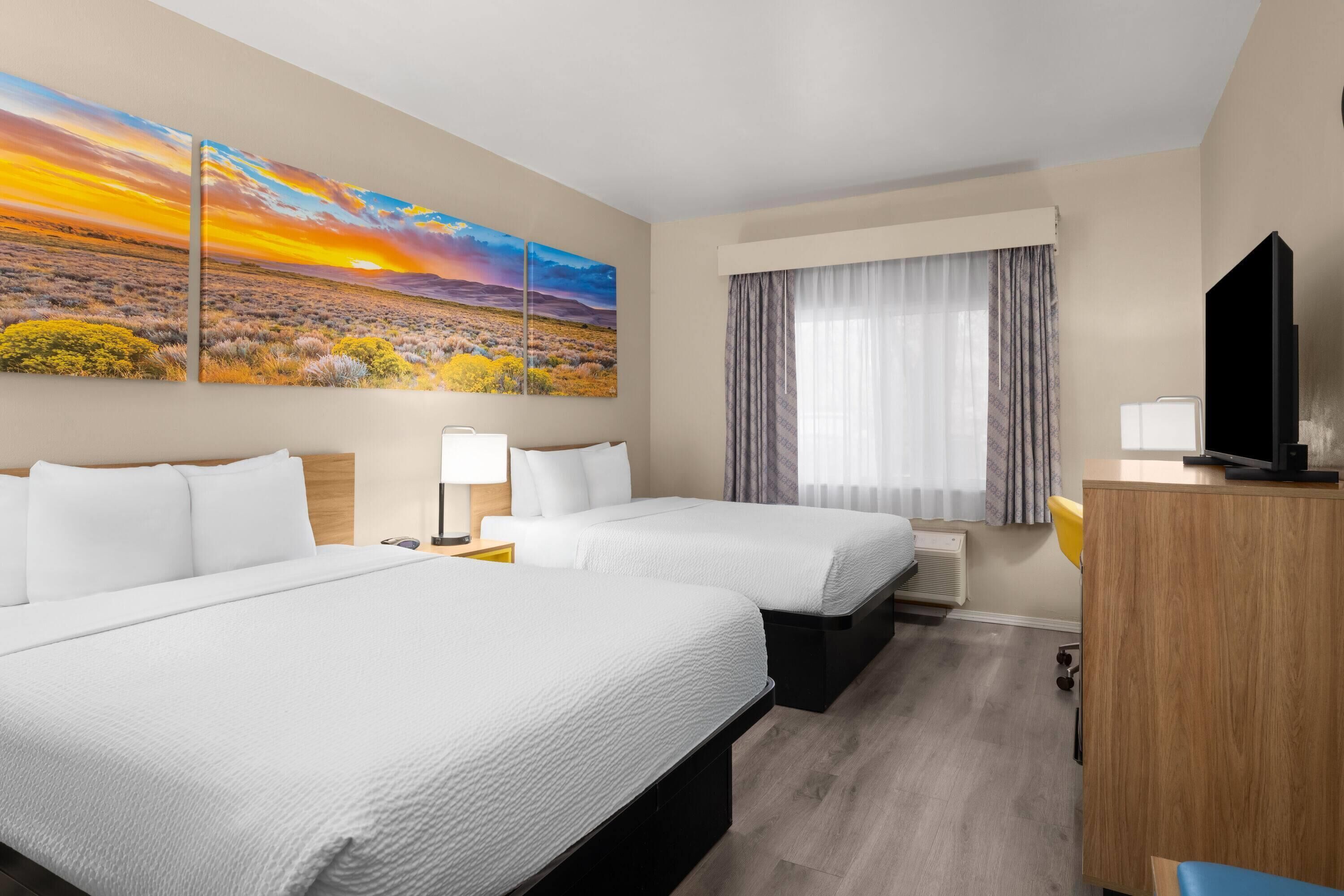 Une chambre d’hôtel avec deux lits, une télévision, un bureau, une lampe et un tableau représentant un paysage au coucher du soleil.