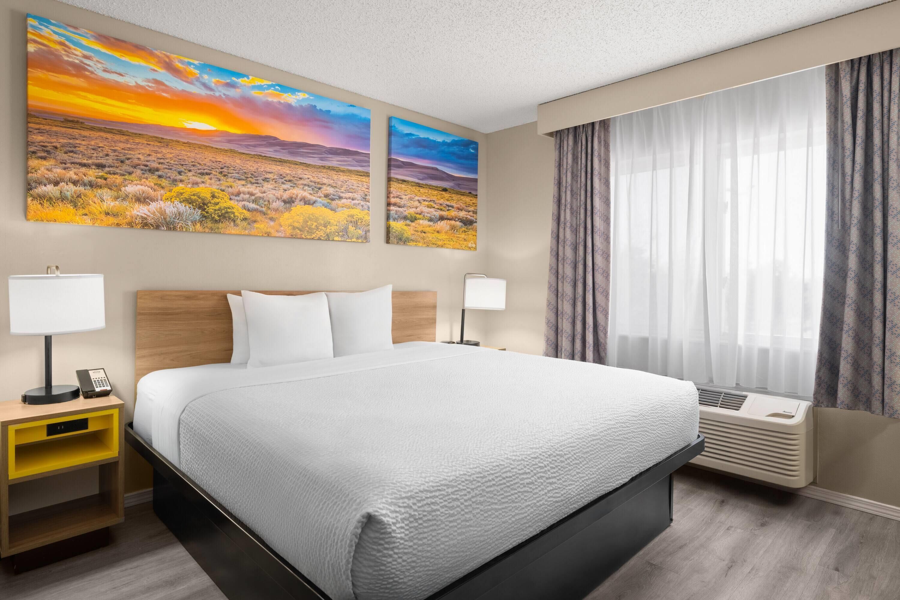Hotelkamer met een bed, twee wanddecoraties, een nachtkastje met lamp, een bijzettafeltje en een raam met gordijnen.