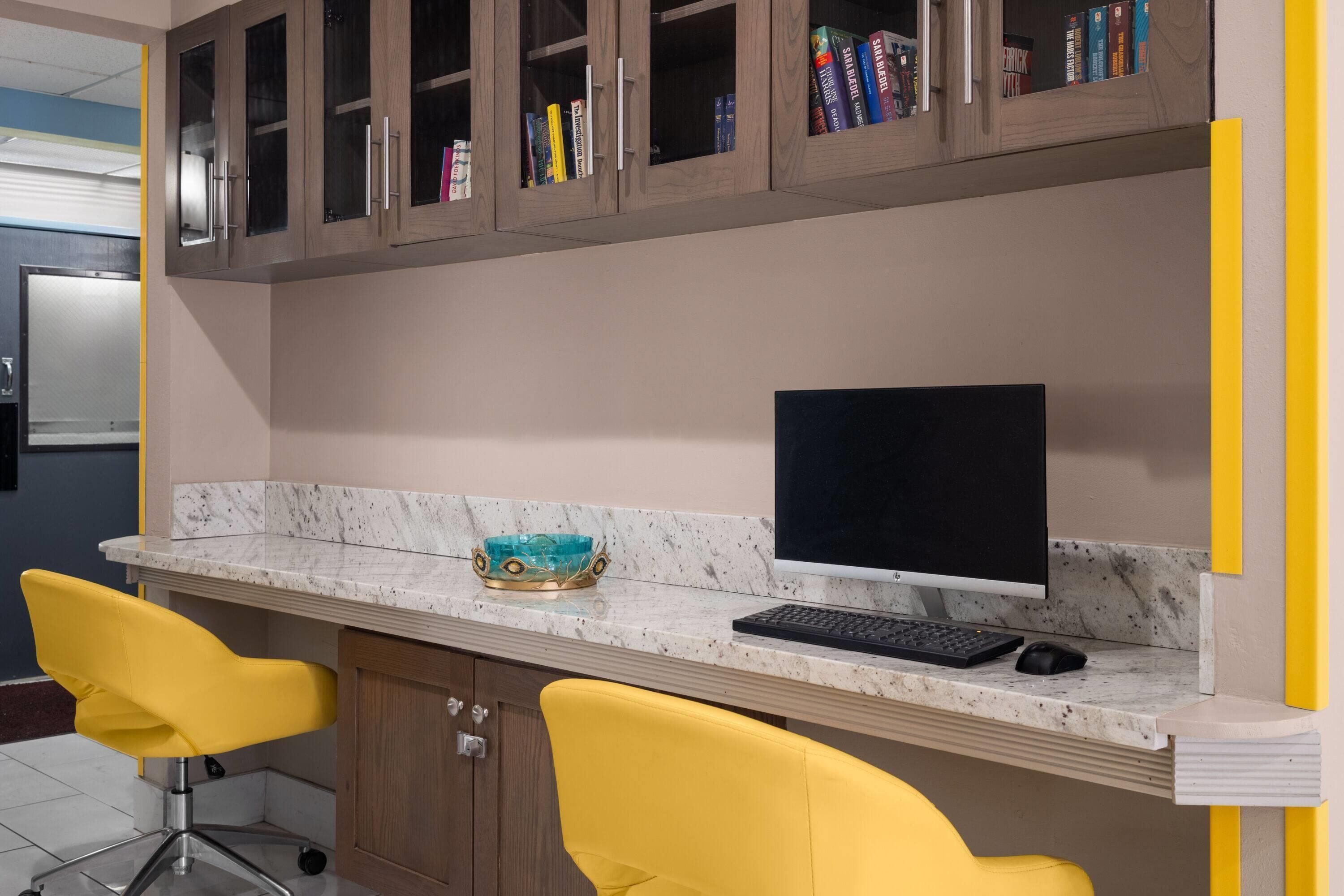 A workspace with a desktop computer, two yellow chairs, a marble countertop, and wooden cabinets with books.