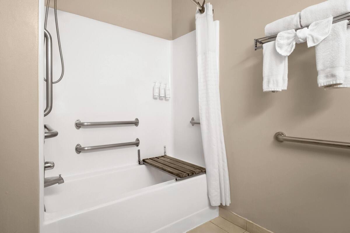 A bathroom with a bathtub, grab bars, a showerhead, a towel rack, and a wooden shelf.
