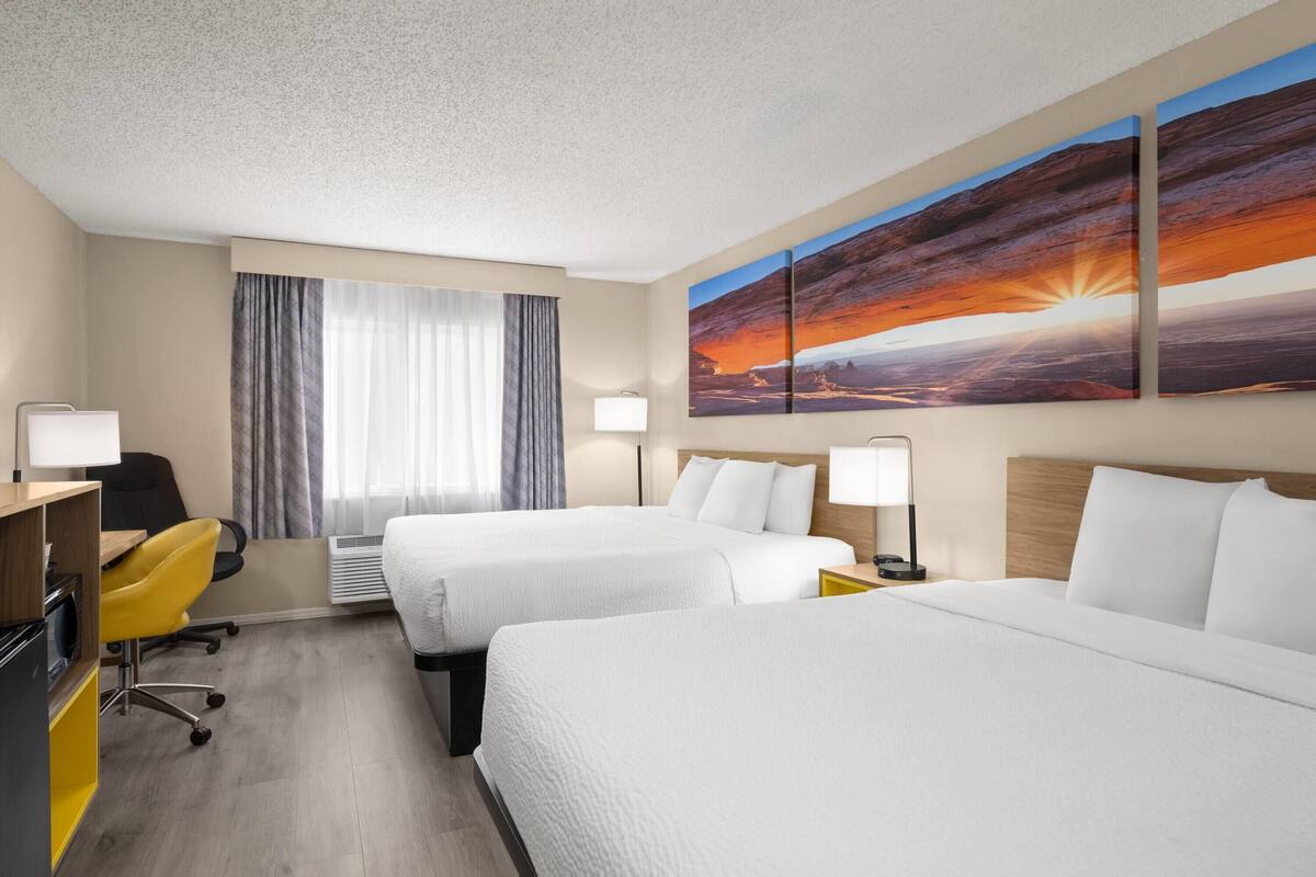 A hotel room with two beds, a desk, a chair, a lamp, and a large painting of a sunset.
