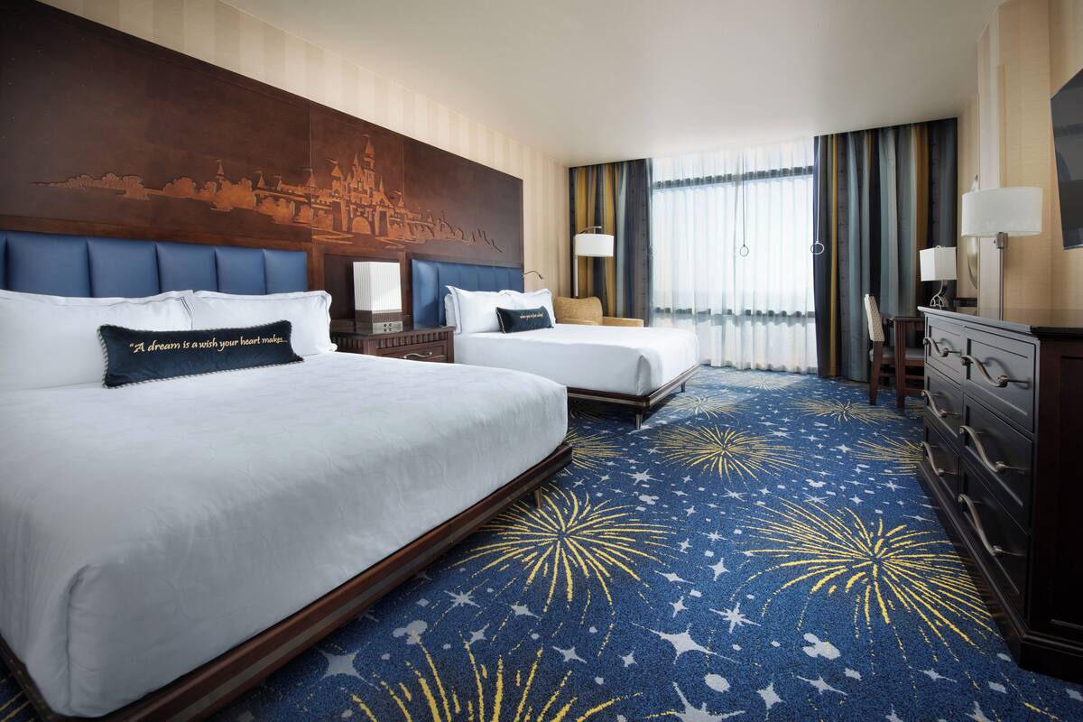 Ein Hotelzimmer mit zwei Betten, einem großen Wandgemälde, einem teppichartigen Boden mit Sternenmuster und einer Kommode mit Lampen.