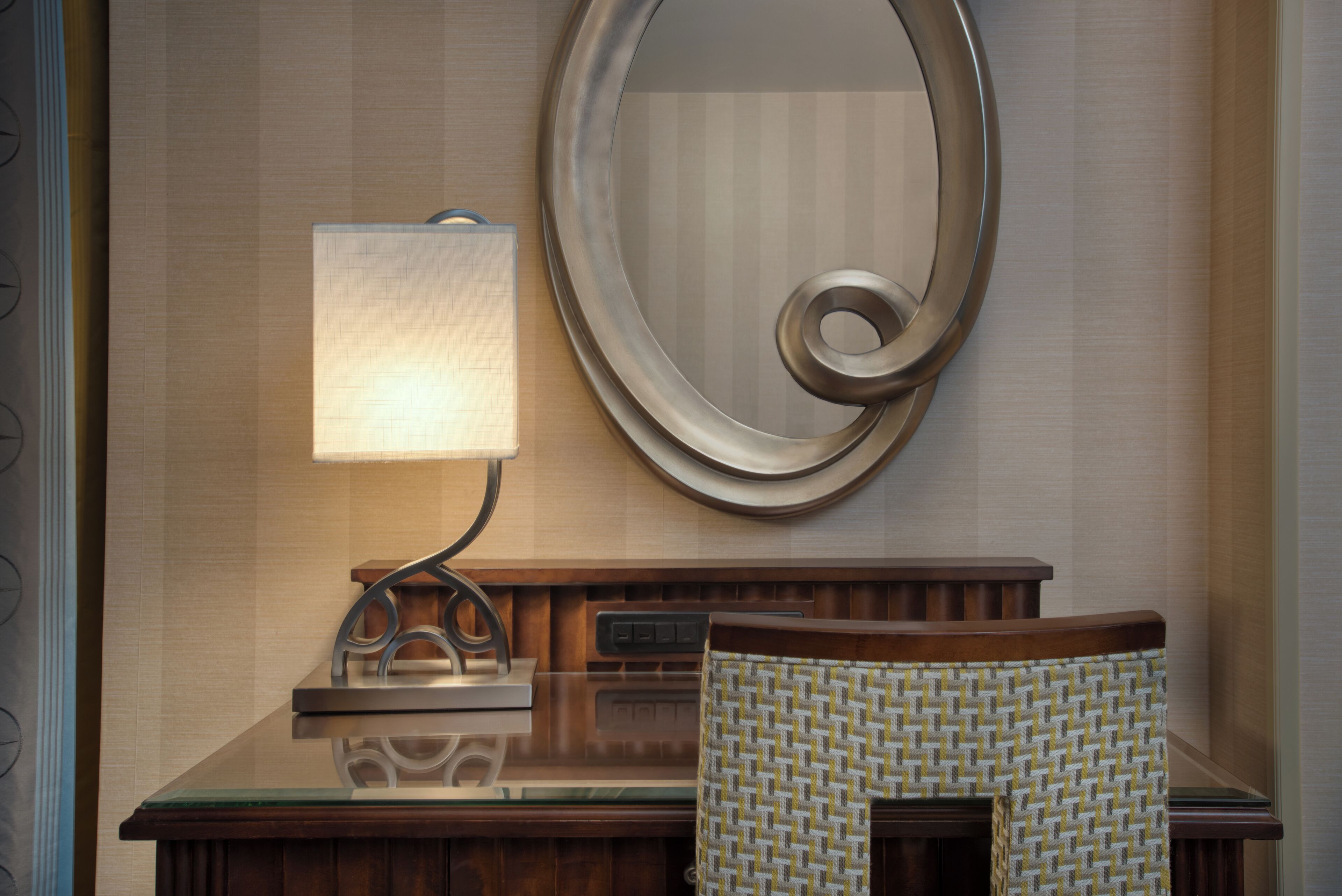 A hotel reception desk with a lamp, a mirror, and a patterned chair.