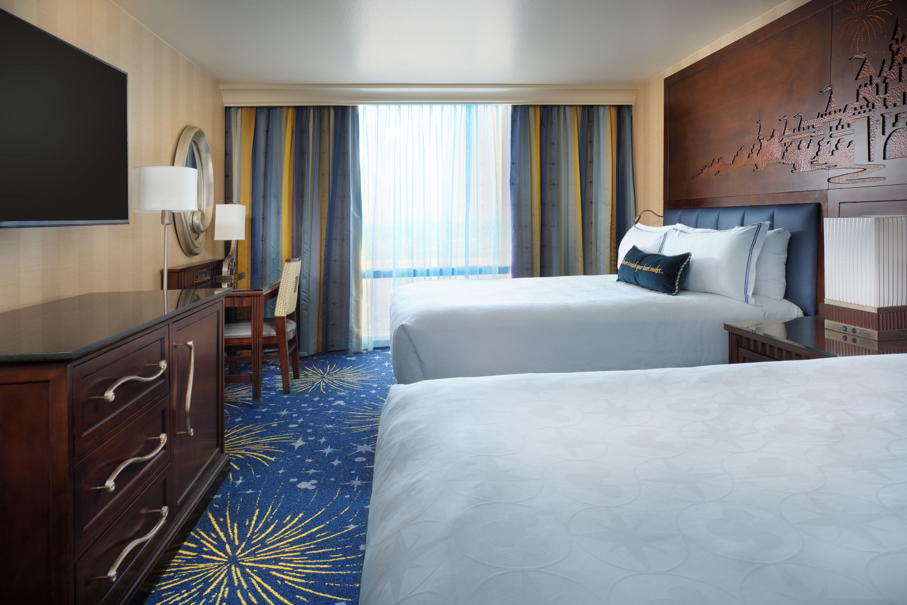A hotel room with two beds, a desk, a chair, a TV, and a large window with curtains.