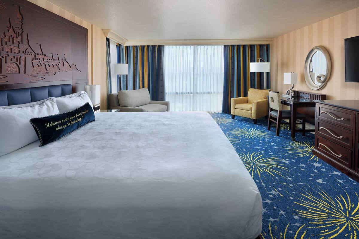 Ein Hotelzimmer mit einem großen Bett, einem Schreibtisch, einem Stuhl und Blick auf die Skyline der Stadt durch das Fenster.