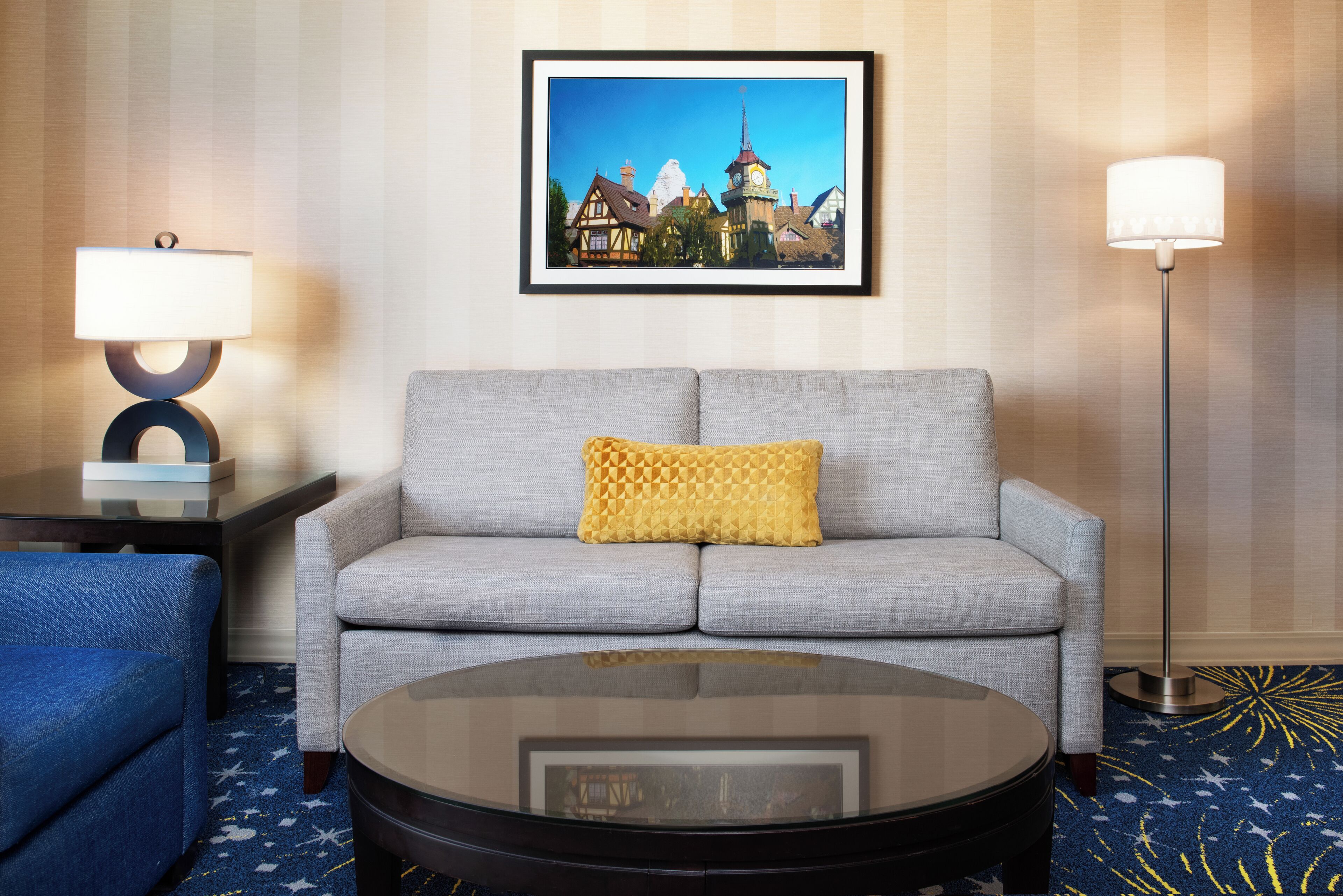 Une chambre d’hôtel avec un canapé gris, une table basse ronde, un fauteuil bleu, un tableau encadré au mur et deux lampadaires.