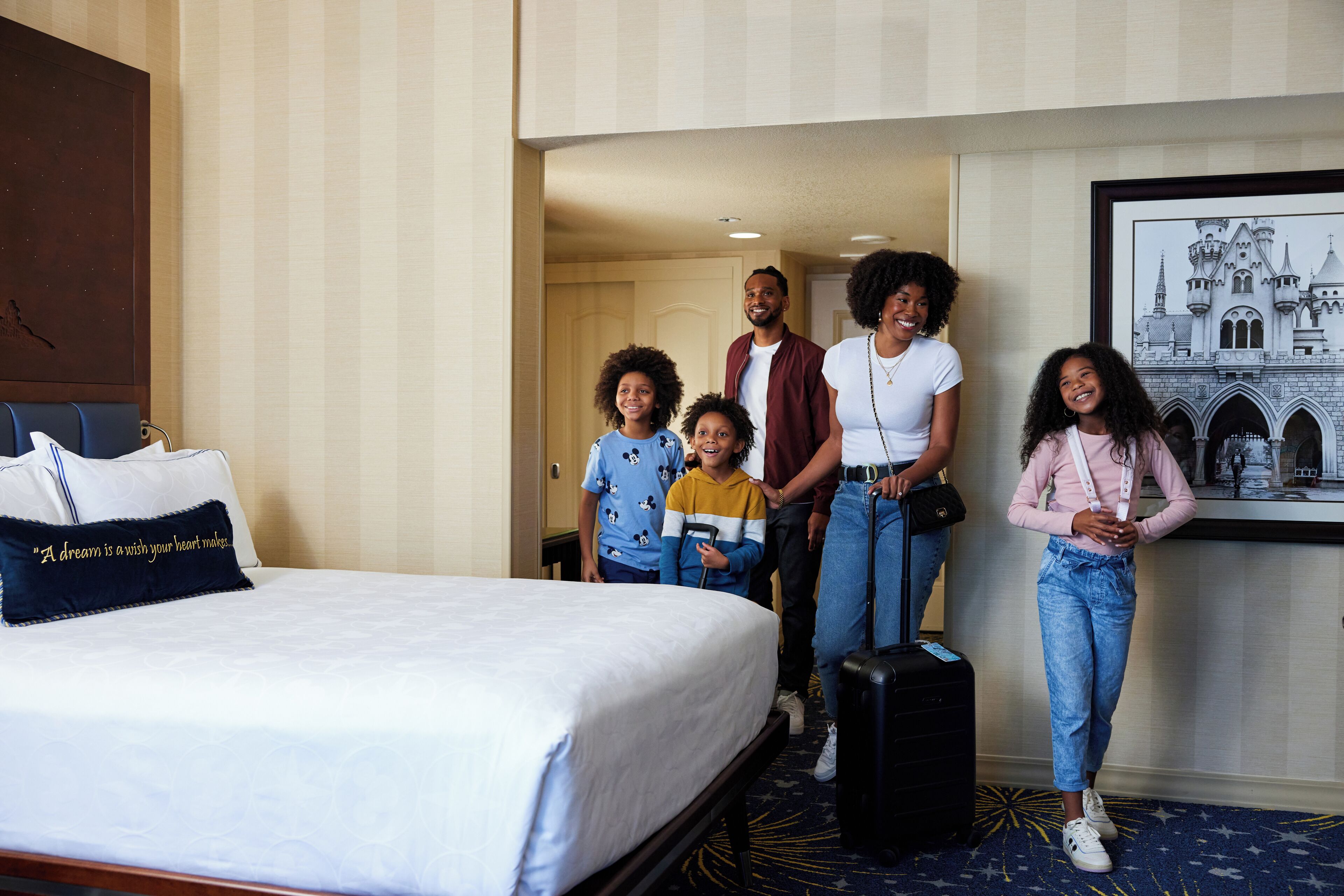 A hotel room with a bed, a pillow with a quote, a framed picture on the wall, and a suitcase on the floor.