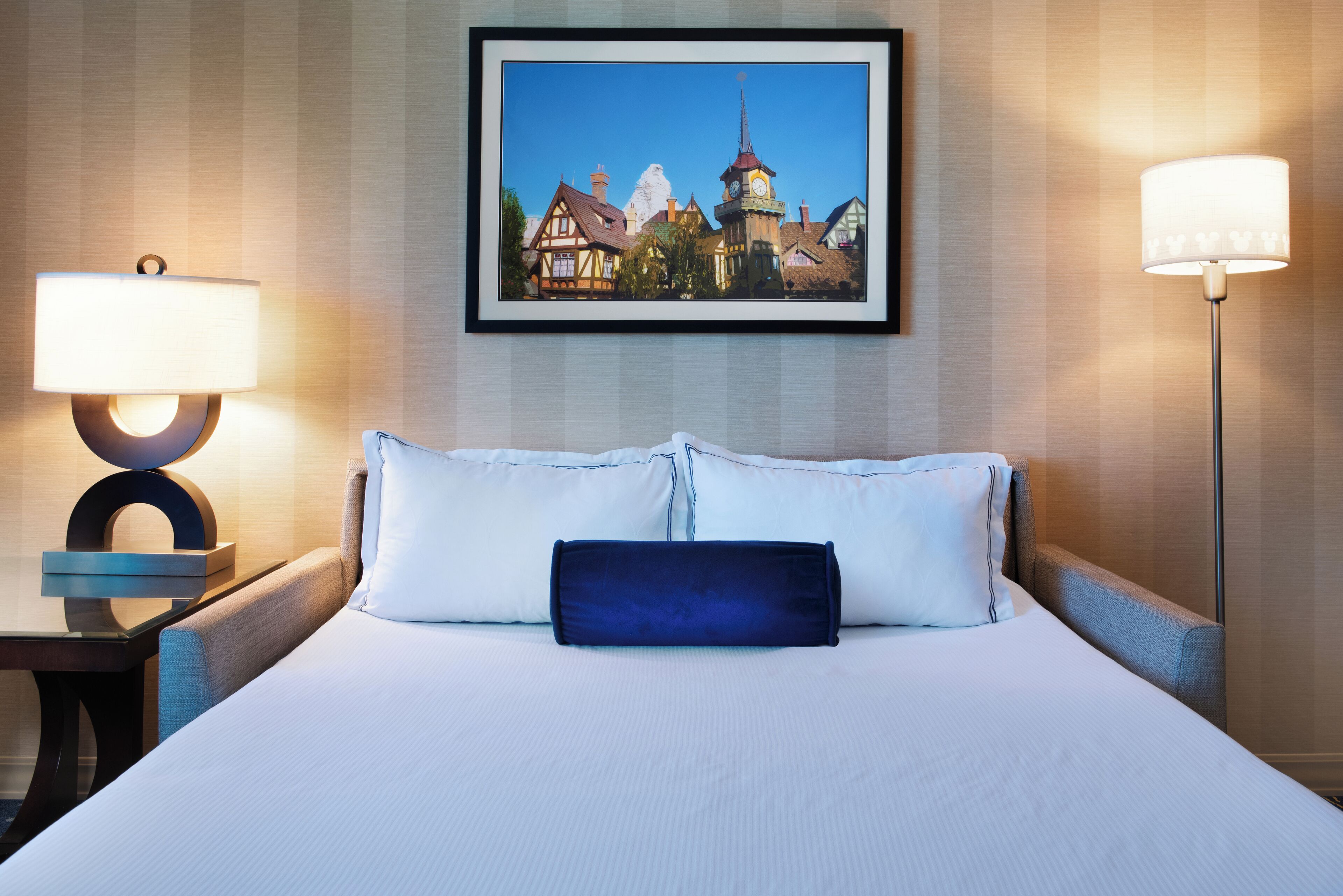 A hotel room with a neatly made bed, two bedside tables with lamps, and a framed picture of a town on the wall.
