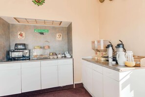 Ein Frühstücksbereich mit Mikrowelle, Toaster und Kaffeemaschine. Unten befinden sich Schränke und ein Schild mit den Frühstückszeiten.