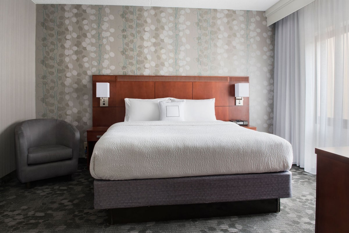 A hotel room with a large bed, a wooden headboard, two bedside lamps, a gray armchair, and a patterned wallpaper.