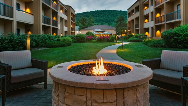 A courtyard with a stone fire pit surrounded by wicker chairs, a pathway leading to a building with multiple balconies, and a lush green lawn. - Courtyard by Marriott Mahwah (Mahwah)