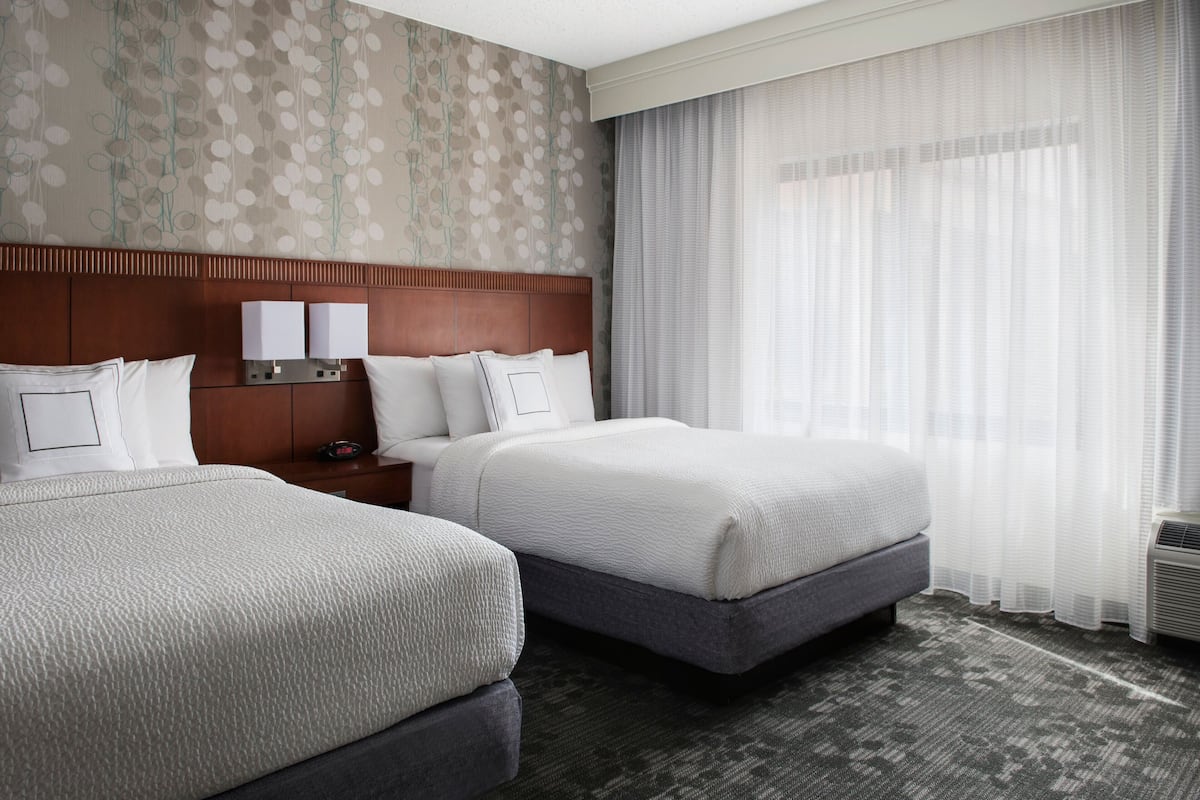 A hotel room with two beds, a wooden headboard, a patterned wall, and a large window with sheer curtains.