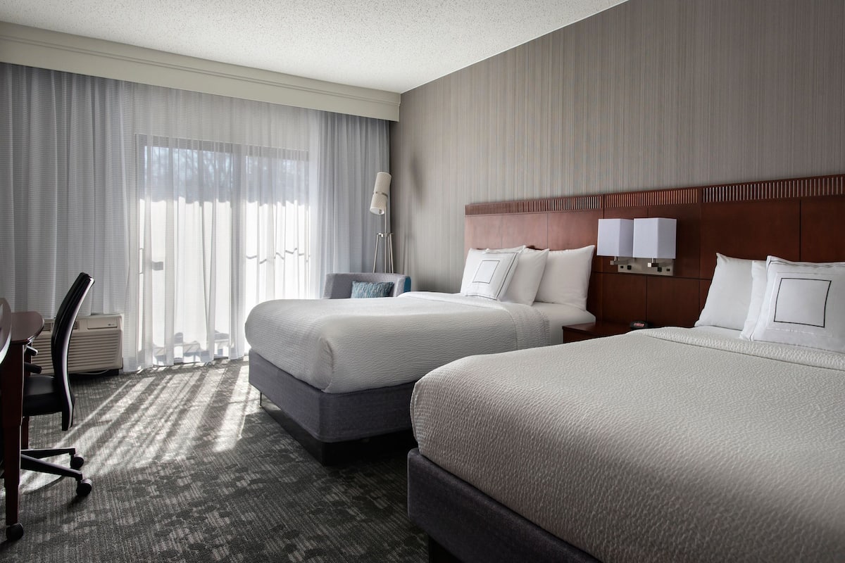 A hotel room with two beds, a desk, a chair, a lamp, and a large window with curtains.