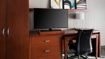 A hotel room with a wooden desk, a black office chair, a flat-screen TV, a lamp, and a set of drawers.