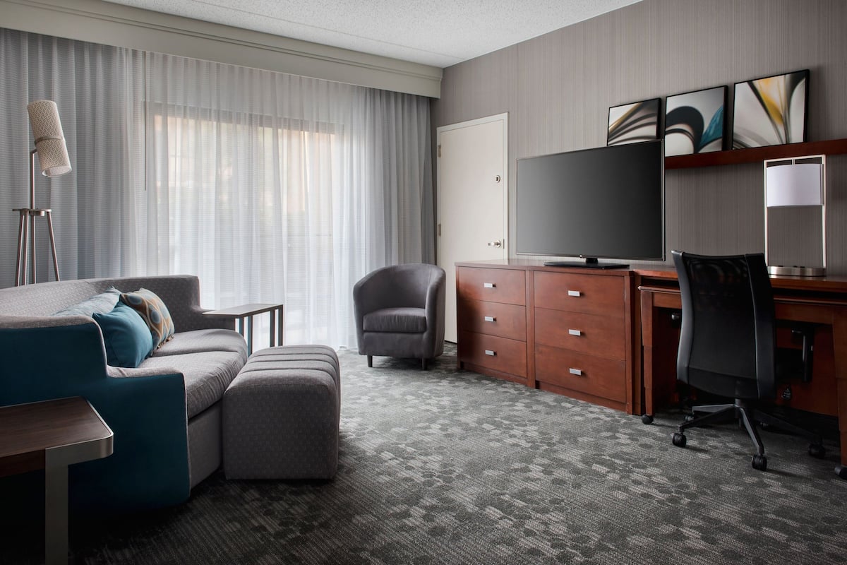 A hotel room with a grey sofa, a small armchair, a desk with a computer, a flat-screen TV, and a large window with sheer curtains.