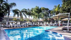 Una piscina con sillas de descanso y sombrillas, rodeada de palmeras y un edificio con tejado de baldosas.