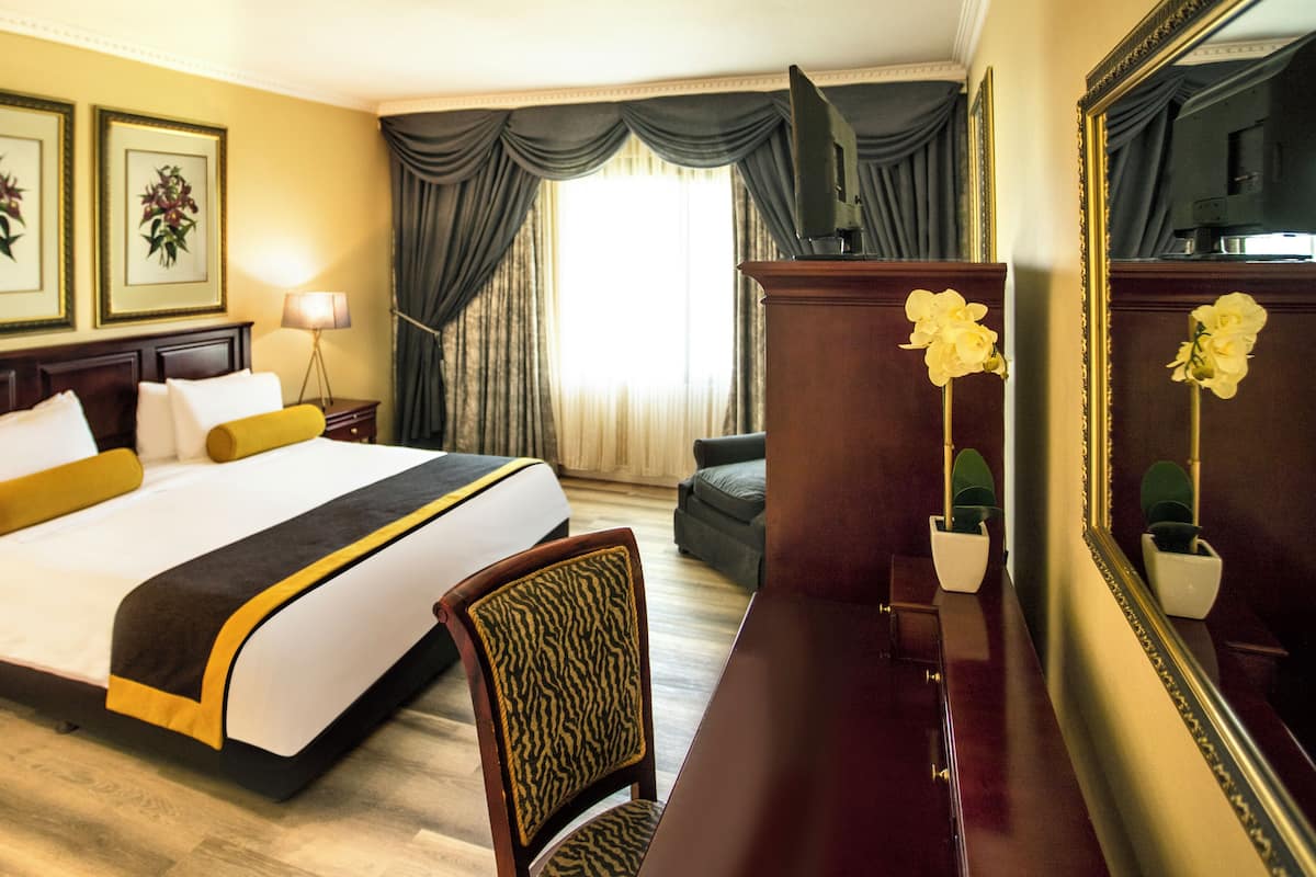 Une chambre d’hôtel comprenant un lit, un bureau, une chaise, une lampe, un miroir et une fenêtre avec des rideaux.