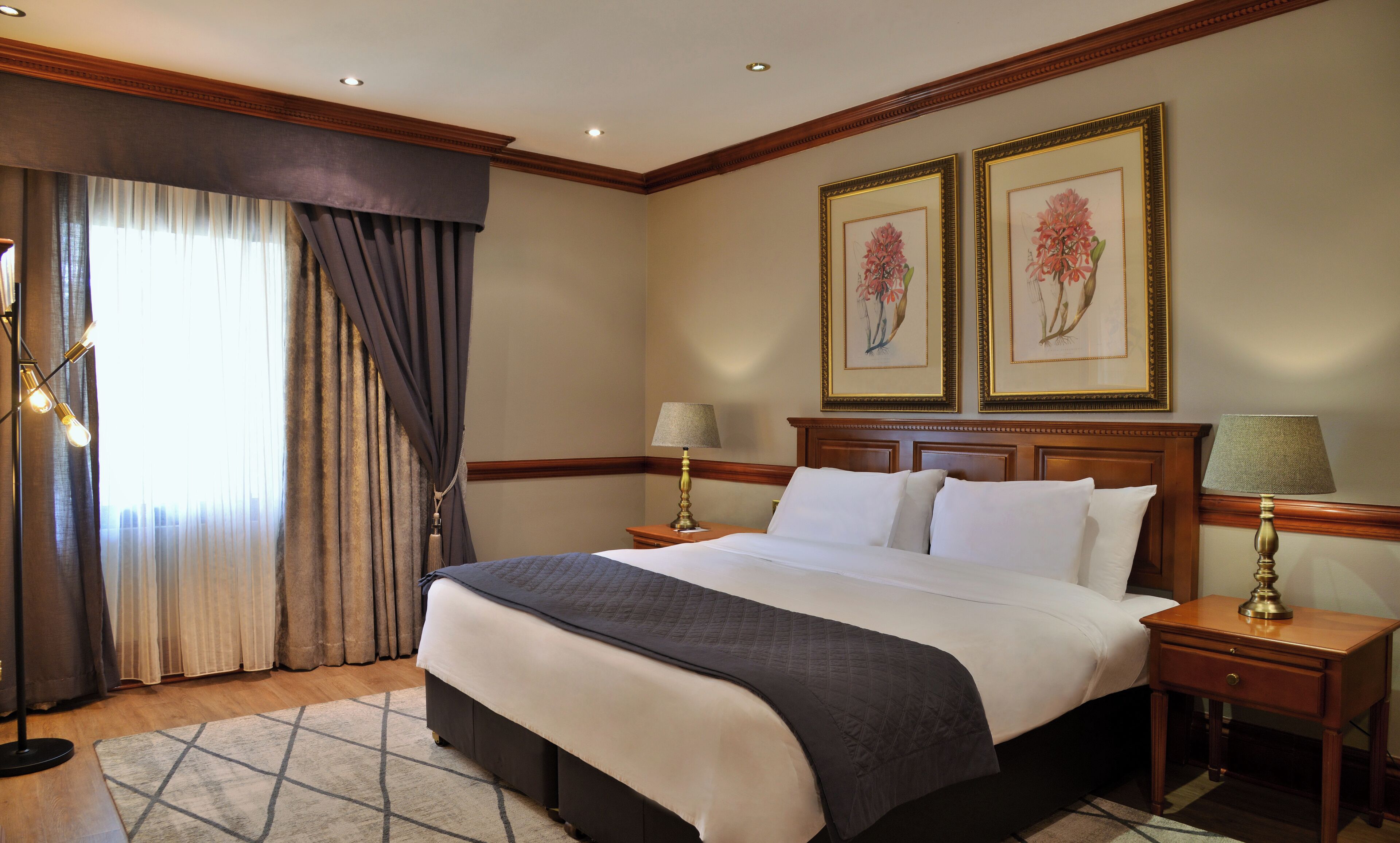 A hotel room with a large bed, two bedside tables with lamps, a window with curtains, and two framed floral paintings on the wall.