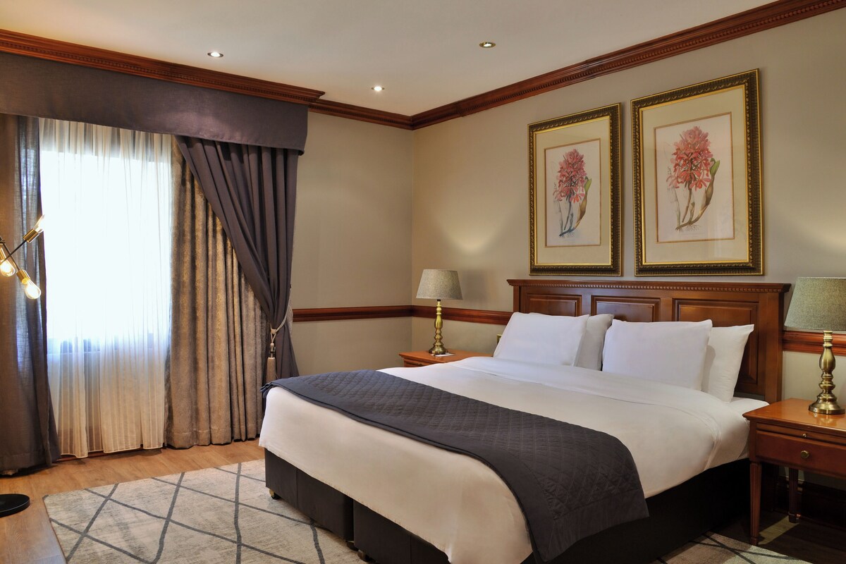 Un cuarto de hotel con una cama grande, dos mesitas de noche con lámparas, un ventanal con cortinas y dos cuadros con motivos florales en la pared.