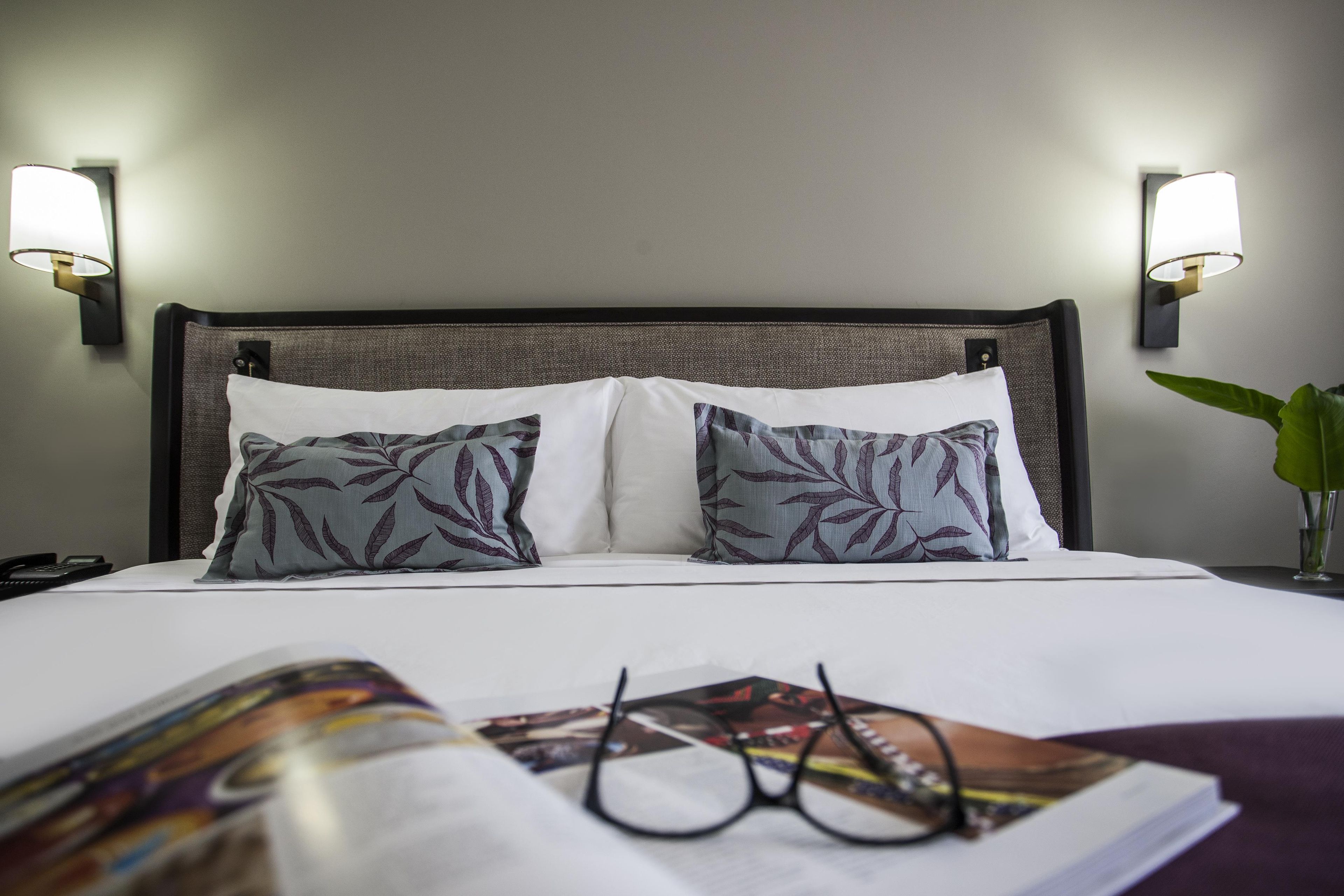 Habitación de hotel con cama, dos almohadas, mesita de noche, lámpara, teléfono y revista.