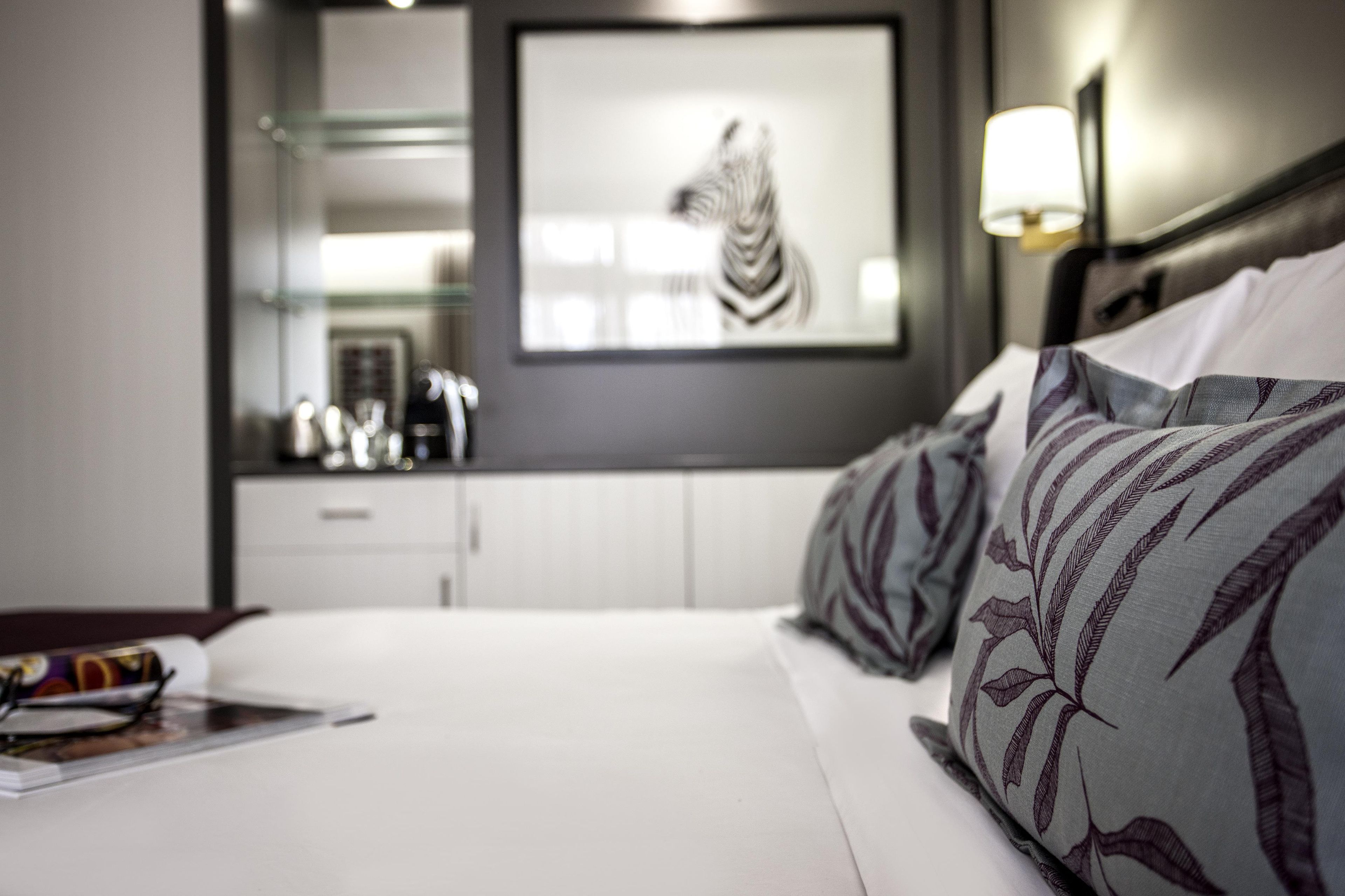 A hotel room with a bed, a nightstand, a mirror, a framed artwork, and a kitchenette area.