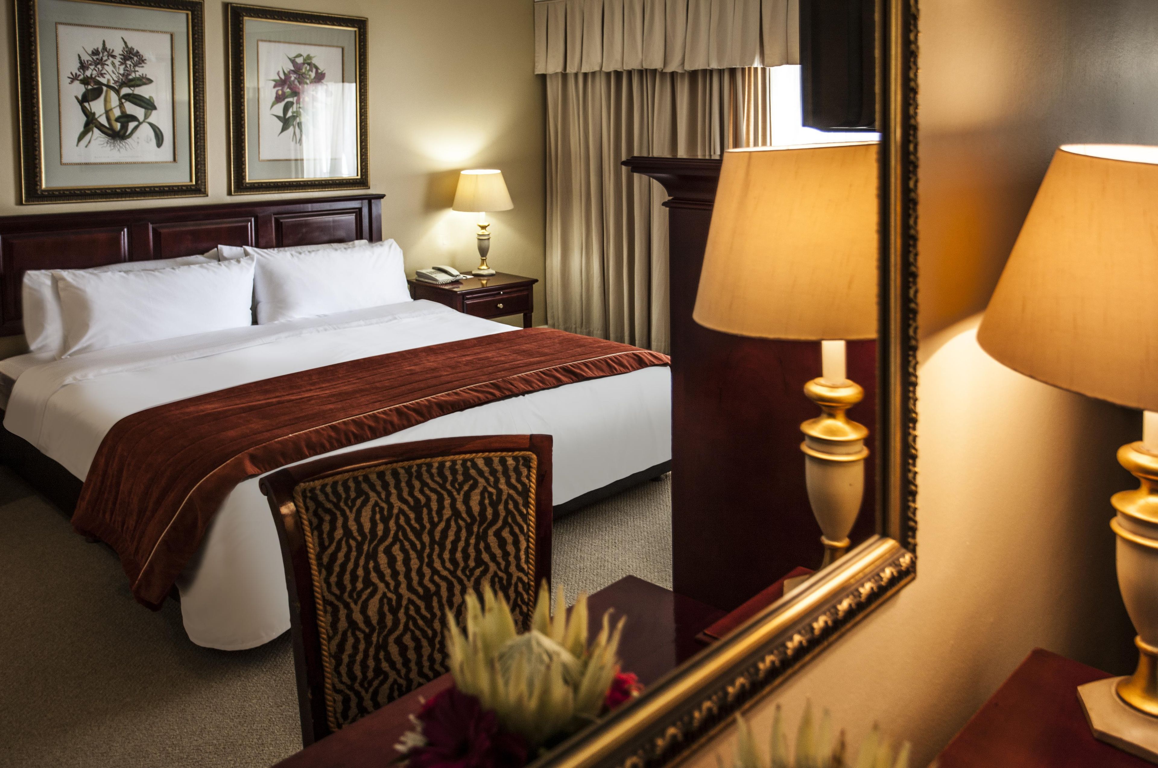 A hotel room with a bed, bedside lamps, a mirror, and framed botanical prints on the wall.