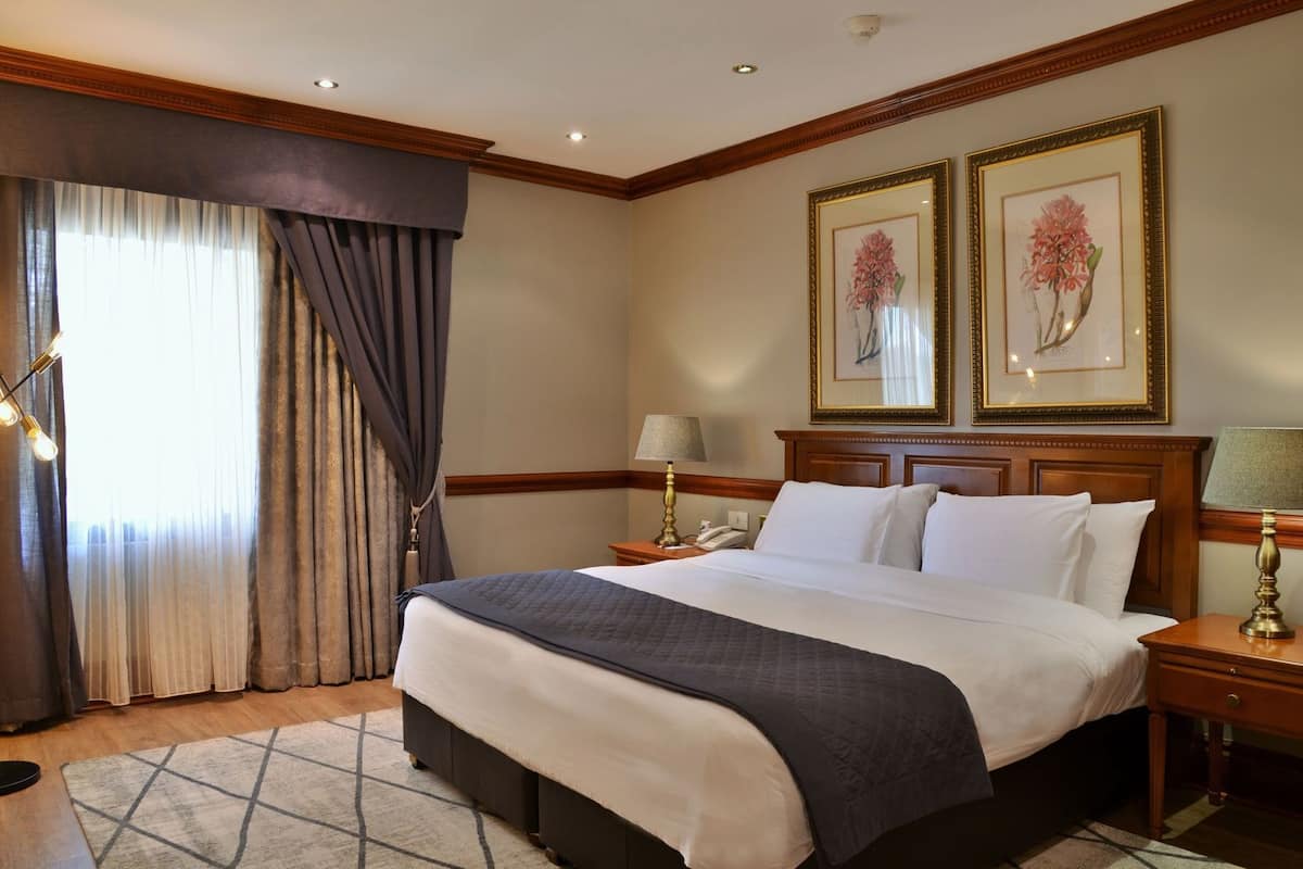 Une chambre d’hôtel avec un grand lit, deux tables de chevet, une lampe sur pied, une fenêtre avec des rideaux et deux tableaux encadrés au mur.