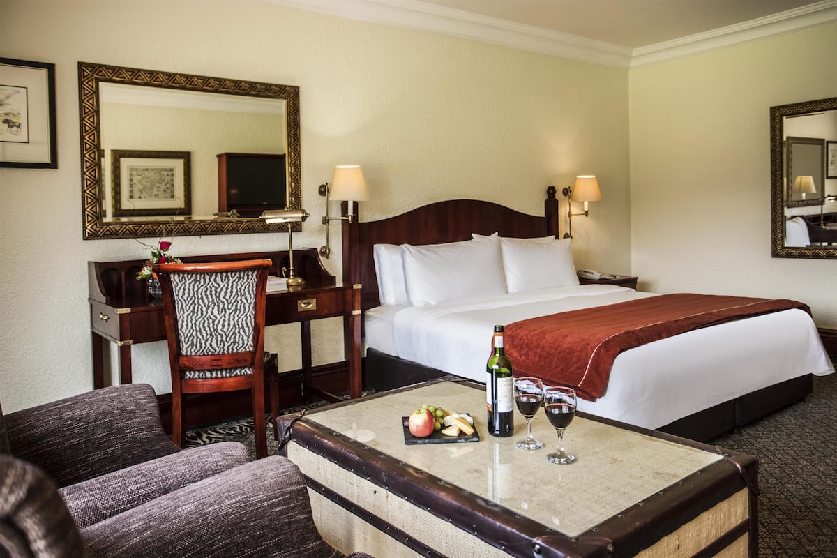 Une chambre d’hôtel avec un lit, un bureau, une chaise, une table avec des fruits et du vin, et un miroir au mur.