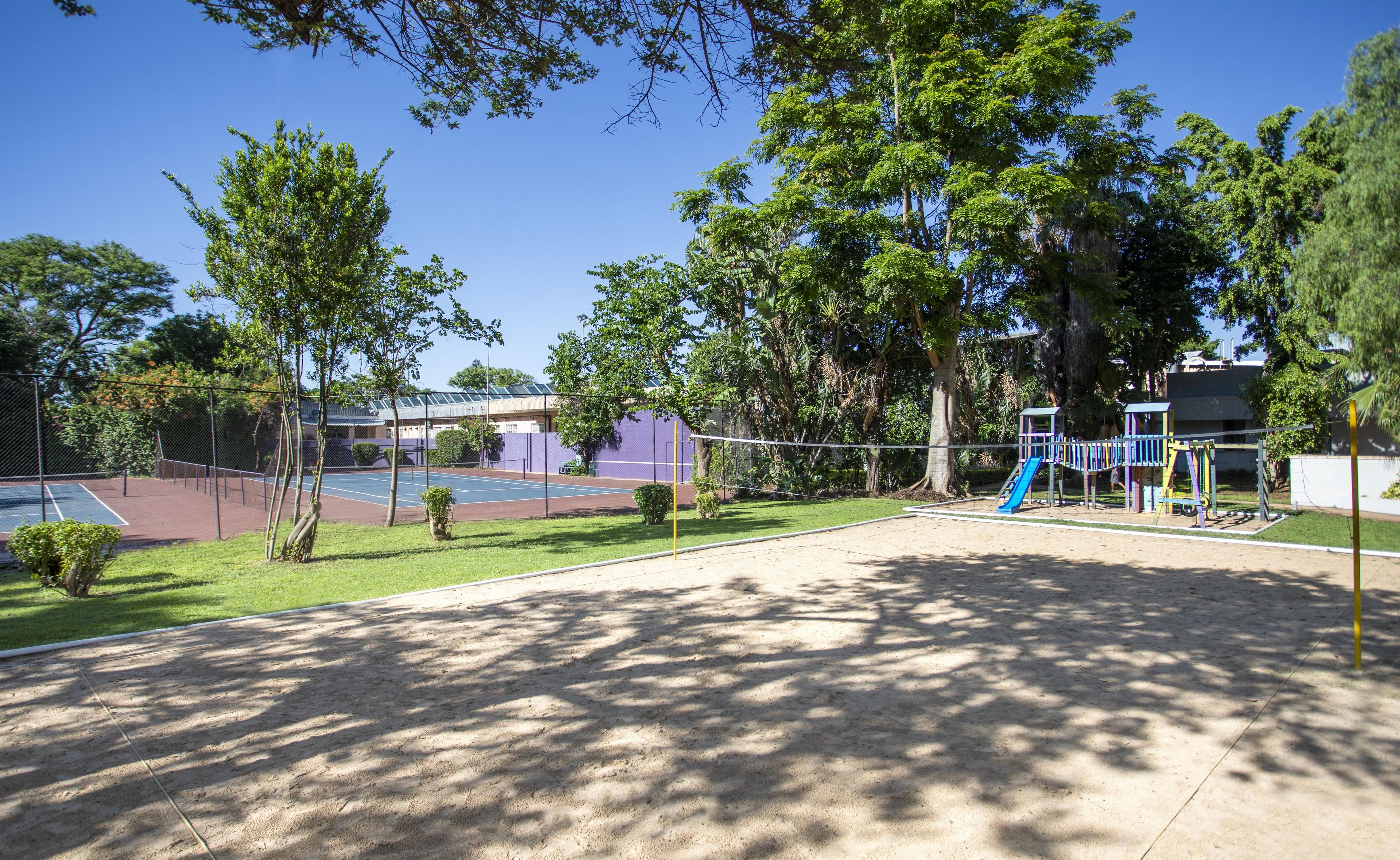 Un court de tennis entouré d’une clôture, avec des arbres et une aire de jeux dotée d’un toboggan.