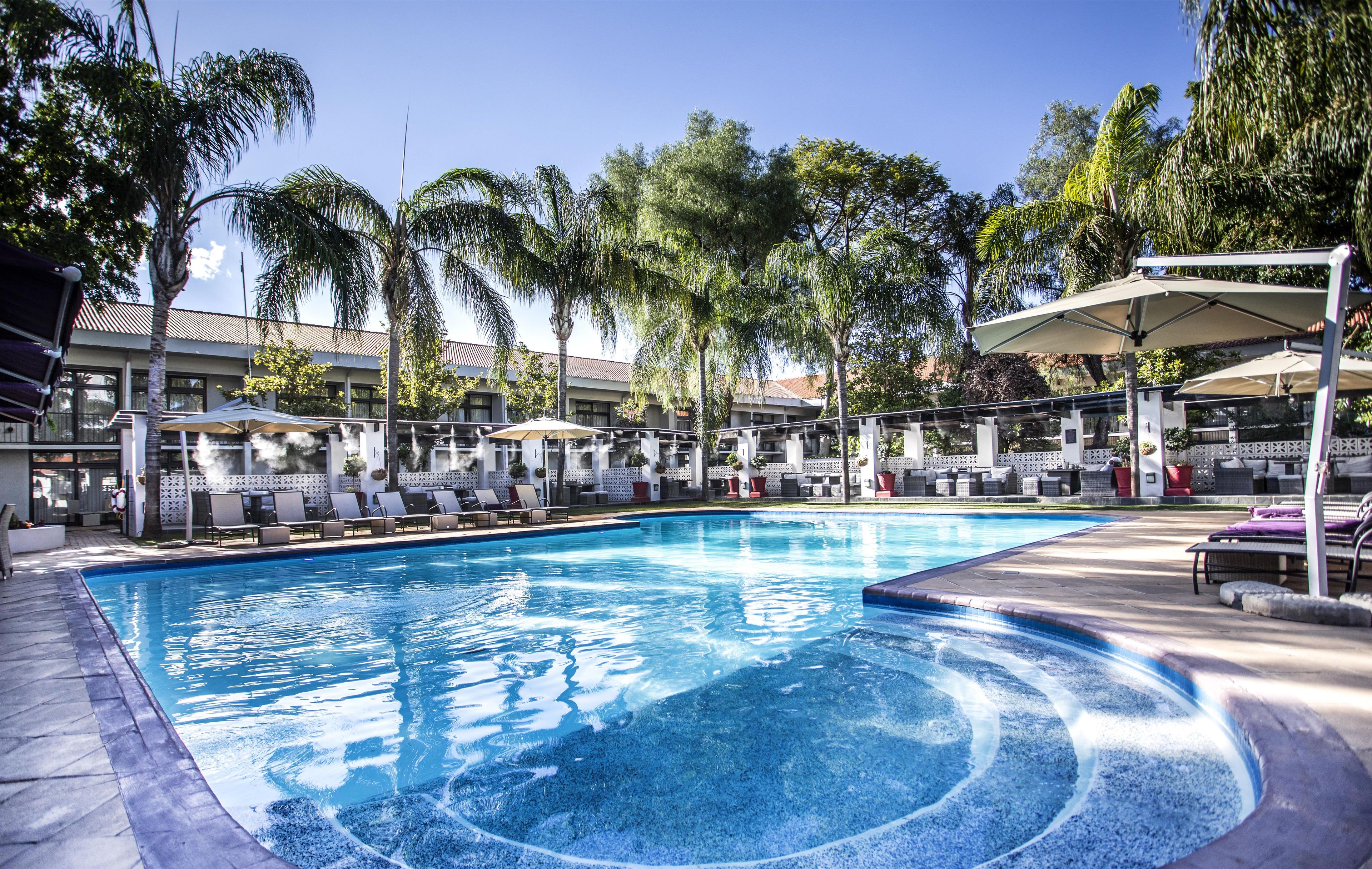 Une piscine entourée de chaises longues et de parasols, bordée de palmiers et d’un bâtiment doté d’un toit en tuiles.