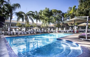 A swimming pool with lounge chairs and umbrellas, surrounded by palm trees and a building with a tiled roof. - Avani Gaborone Resort & Casino (Gaborone)