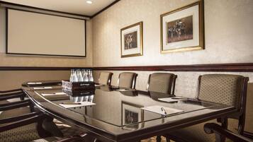 Un salón de conferencias con una mesa de vidrio, sillas, una pantalla de proyector y cuadros en la pared.